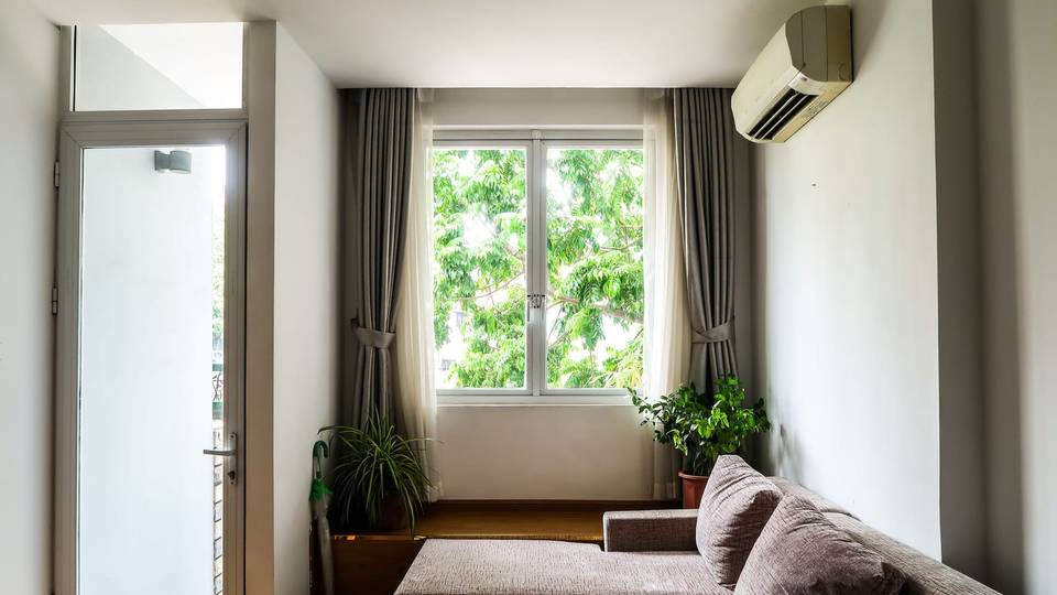 A bright room with a window, grey curtains and an older wall-mounted split AC due for cleaning